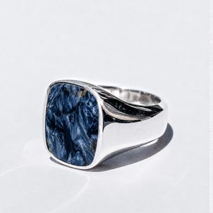 Puede incluir: Un anillo de sello de plata con una incrustación de piedra pietersita azul pulida de forma cuadrada. La piedra muestra un patrón arremolinado y chatoyante de vetas azul marino profundo y azul claro, engastado en una banda metálica lisa.