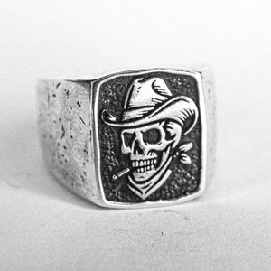 May include: A silver ring with a detailed skull design, wearing a cowboy hat and bandana, and smoking a cigarette. The skull is set within a rectangular frame. The ring has a textured, worn appearance, with a vintage aesthetic.