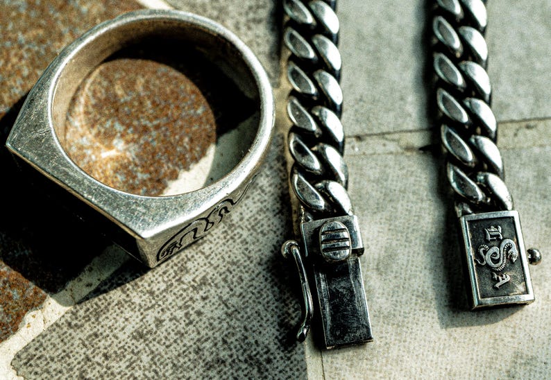 May include: A silver ring with an intricate design and a silver chain bracelet with a clasp that has a snake design. The bracelet has the text "The Serpent" on the clasp.
