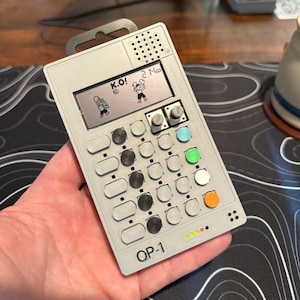 Puede incluir: Un dispositivo electrónico gris portátil con una pantalla que muestra un juego de boxeo con las palabras "K.O.!" y "2:14". El dispositivo tiene varios botones de diferentes colores y un lazo de metal en la parte superior. El texto "OP-1" está impreso en la parte inferior.