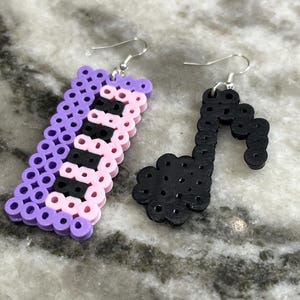 Può includere: Un paio di orecchini realizzati con perline di plastica colorate. Un orecchino ha la forma di una nota musicale e l'altro ha la forma di una tastiera di pianoforte.