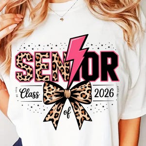 Puede incluir: Camiseta blanca con un diseño "SENIOR" con estampado de leopardo, un rayo rosa y un lazo con estampado de leopardo. También incluye el texto "Class of 2026". Es una prenda con temática de graduación.