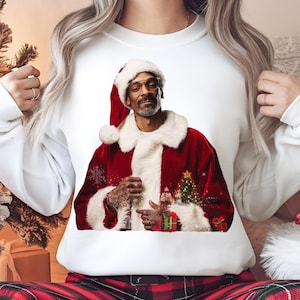 May include: A white sweatshirt featuring a printed design of a person dressed as Santa Claus. The person is wearing a red and white Santa suit, a Santa hat, and sunglasses. The graphic is surrounded by snowflakes and a small Christmas tree.