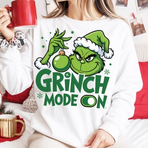 Op de afbeelding: Witte sweatshirt met een groene Grinch-afbeelding die een groene kerstbal vasthoudt. De tekst "GRINCH MODE ON" staat eronder gedrukt. Het ontwerp bevat groene sneeuwvlokken en een groen-witte hoed.