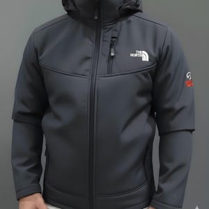 以下が含まれることがあります： フードとフルレングスのジッパーが付いたチャコールグレーのジャケット。胸には白い「THE NORTH FACE」のロゴ、袖には赤い「SUMMIT SERIES」のロゴが入っています。ジャケットは洗練されたモダンなデザインです。