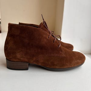 K&ouml;nnte beinhalten: Braune Wildleder-Ankle Boots mit Schn&uuml;rung. Die Stiefel haben einen niedrigen Absatz und eine runde Spitze. Die Farbe ist ein warmes Braun, und die Wildlederstruktur ist sichtbar. Diese Stiefel eignen sich f&uuml;r den Alltag.
