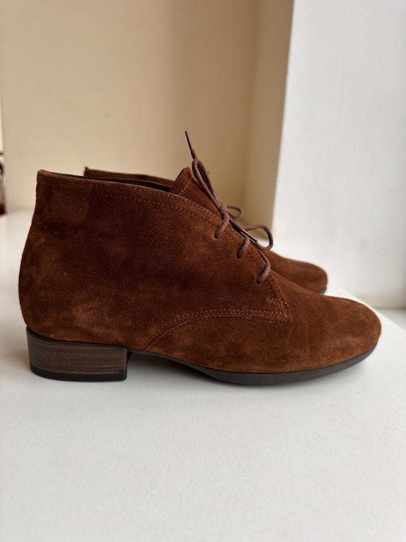 K&ouml;nnte beinhalten: Braune Wildleder-Ankle Boots mit Schn&uuml;rung. Die Stiefel haben einen niedrigen Absatz und eine runde Spitze. Die Farbe ist ein warmes Braun, und die Wildlederstruktur ist sichtbar. Diese Stiefel eignen sich f&uuml;r den Alltag.