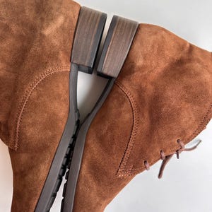 K&ouml;nnte beinhalten: Ein Paar braune Wildlederstiefel mit niedrigem Absatz. Die Stiefel haben ein Schn&uuml;rdesign und eine strukturierte Sohle. Die Farbe ist ein warmes, erdiges Braun, und die Stiefel scheinen ungetragen zu sein.
