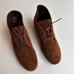 K&ouml;nnte beinhalten: Ein Paar braune Wildleder-Ankle-Boots mit passenden Schn&uuml;rsenkeln. Die Stiefel haben eine runde Spitze und einen kleinen Absatz. Die Schn&uuml;rsenkel sind kreuzweise gebunden. Die Stiefel stehen vor einem wei&szlig;en Hintergrund.