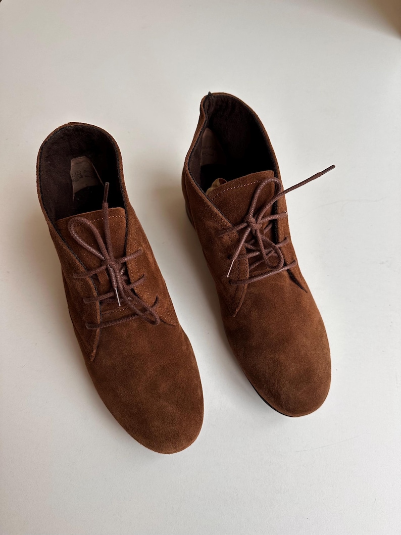 K&ouml;nnte beinhalten: Ein Paar braune Wildleder-Ankle-Boots mit passenden Schn&uuml;rsenkeln. Die Stiefel haben eine runde Spitze und einen kleinen Absatz. Die Schn&uuml;rsenkel sind kreuzweise gebunden. Die Stiefel stehen vor einem wei&szlig;en Hintergrund.