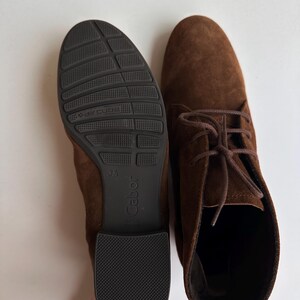 K&ouml;nnte beinhalten: Ein Paar braune Wildleder-Ankle Boots mit Schn&uuml;rsenkeln. Einer der Stiefel zeigt die Sohle, mit einer schwarzen Gummisohle mit dem Markennamen "Gabor" und "aircube". Der andere Stiefel wird von oben mit gebundenen Schn&uuml;rsenkeln gezeigt.
