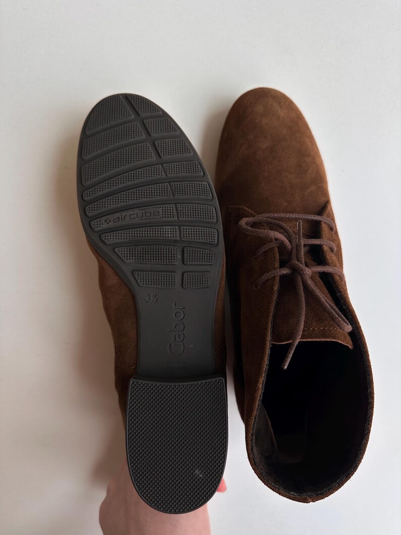 K&ouml;nnte beinhalten: Ein Paar braune Wildleder-Ankle Boots mit Schn&uuml;rsenkeln. Einer der Stiefel zeigt die Sohle, mit einer schwarzen Gummisohle mit dem Markennamen "Gabor" und "aircube". Der andere Stiefel wird von oben mit gebundenen Schn&uuml;rsenkeln gezeigt.