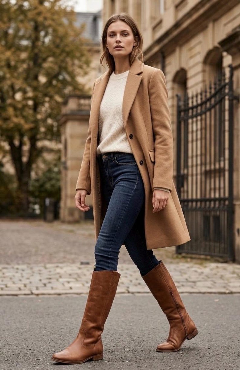 Pu&ograve; includere: Una donna indossa un cappotto color cammello, un maglione color crema e jeans blu scuro. Indossa anche stivali alti al ginocchio in pelle marrone. Lo sfondo include un edificio e un cancello in ferro battuto.