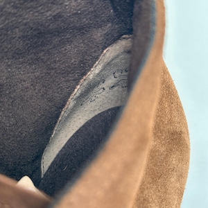 K&ouml;nnte beinhalten: Nahaufnahme des Inneren eines braunen Wildlederstiefels. Der Innenraum hat ein dunkelbraunes, weiches Futter. Die Innensohle ist heller mit einem sichtbaren Logo. Die Au&szlig;enseite des Stiefels ist hellbraunes Wildleder.