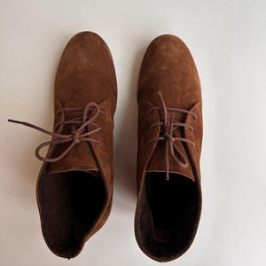 K&ouml;nnte beinhalten: Ein Paar braune Wildleder-Ankle Boots mit passenden Schn&uuml;rsenkeln. Die Stiefel haben ein klassisches Design mit runder Spitze und kleinem Absatz. Die Schuhe werden auf einer wei&szlig;en Oberfl&auml;che pr&auml;sentiert.