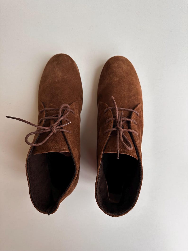 K&ouml;nnte beinhalten: Ein Paar braune Wildleder-Ankle Boots mit passenden Schn&uuml;rsenkeln. Die Stiefel haben ein klassisches Design mit runder Spitze und kleinem Absatz. Die Schuhe werden auf einer wei&szlig;en Oberfl&auml;che pr&auml;sentiert.