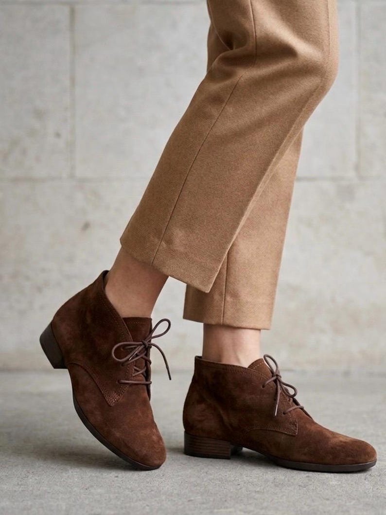 K&ouml;nnte beinhalten: Braune Wildleder-Ankle Boots mit Schn&uuml;rung. Die Stiefel haben einen kleinen Absatz und werden mit einer beigefarbenen Hose kombiniert. Die Schuhe sind ein klassischer Stil, der f&uuml;r verschiedene Anl&auml;sse geeignet ist.