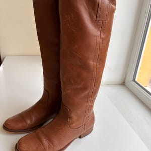 Pu&ograve; includere: Un paio di stivali alti in pelle marrone. Gli stivali hanno un design classico con gambale alto, punta arrotondata e tacco basso. La pelle ha un aspetto leggermente invecchiato. Gli stivali misurano circa 40 cm di altezza.