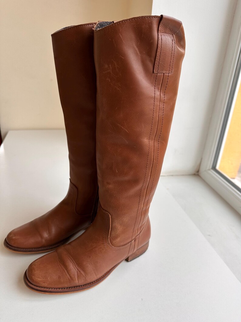 Pu&ograve; includere: Un paio di stivali alti in pelle marrone. Gli stivali hanno un design classico con gambale alto, punta arrotondata e tacco basso. La pelle ha un aspetto leggermente invecchiato. Gli stivali misurano circa 40 cm di altezza.