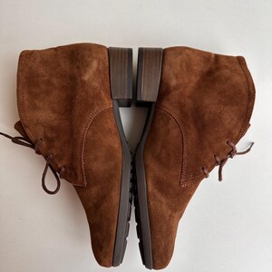 K&ouml;nnte beinhalten: Ein Paar braune Wildleder-Ankle Boots mit Schn&uuml;rung. Die Stiefel haben einen niedrigen Blockabsatz und eine runde Spitze. Das Wildleder hat eine weiche, strukturierte Optik. Die Stiefel werden auf einer wei&szlig;en Oberfl&auml;che pr&auml;sentiert.