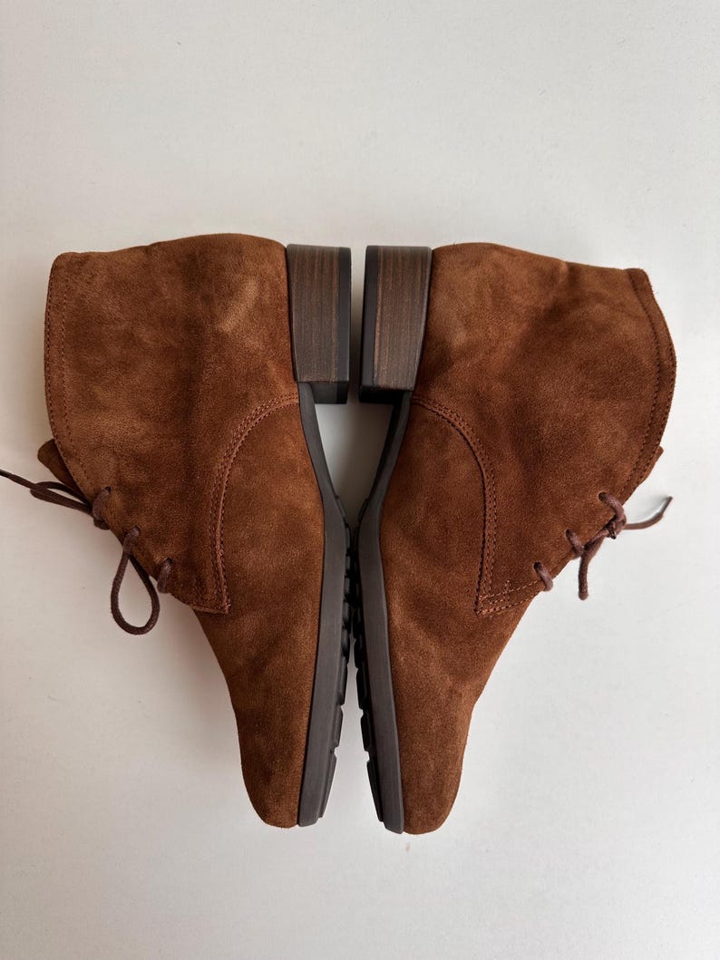 K&ouml;nnte beinhalten: Ein Paar braune Wildleder-Ankle Boots mit Schn&uuml;rung. Die Stiefel haben einen niedrigen Blockabsatz und eine runde Spitze. Das Wildleder hat eine weiche, strukturierte Optik. Die Stiefel werden auf einer wei&szlig;en Oberfl&auml;che pr&auml;sentiert.