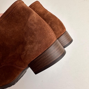 K&ouml;nnte beinhalten: Nahaufnahme von zwei braunen Wildleder-Ankle-Boots. Die Stiefel haben einen niedrigen, gestapelten Holzabsatz und ein weiches, strukturiertes Obermaterial. Die Farbe ist ein warmes, r&ouml;tliches Braun.