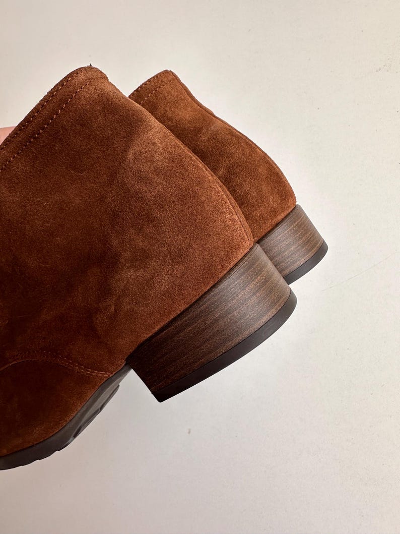 K&ouml;nnte beinhalten: Nahaufnahme von zwei braunen Wildleder-Ankle-Boots. Die Stiefel haben einen niedrigen, gestapelten Holzabsatz und ein weiches, strukturiertes Obermaterial. Die Farbe ist ein warmes, r&ouml;tliches Braun.
