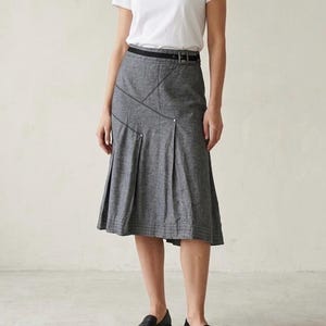 Peut inclure: Jupe midi grise avec une ceinture noire et des coutures décoratives. La jupe présente des plis et une forme légèrement évasée. Le modèle porte un t-shirt blanc et des chaussures noires.