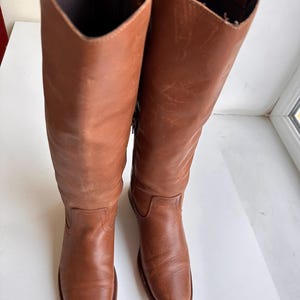 Pu&ograve; includere: Un paio di stivali alti in pelle marrone. Gli stivali presentano un design classico, con una punta arrotondata e un gambale alto. La pelle ha un aspetto leggermente invecchiato, che ne aumenta il fascino vintage. Gli stivali sono esposti su una superficie bianca.