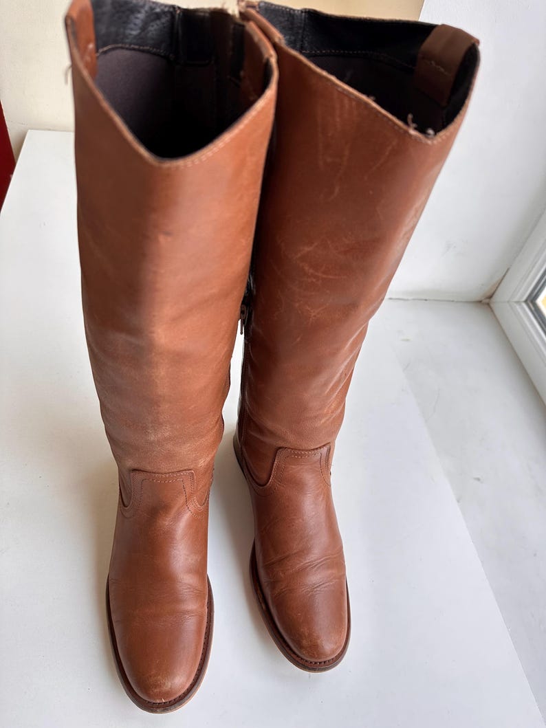 Pu&ograve; includere: Un paio di stivali alti in pelle marrone. Gli stivali presentano un design classico, con una punta arrotondata e un gambale alto. La pelle ha un aspetto leggermente invecchiato, che ne aumenta il fascino vintage. Gli stivali sono esposti su una superficie bianca.