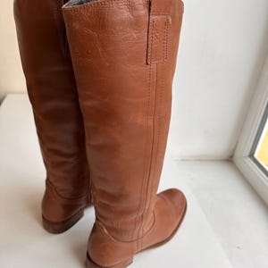 Pu&ograve; includere: Un paio di stivali alti in pelle marrone. Gli stivali hanno un design al ginocchio con punta arrotondata e tacco basso. La pelle ha una consistenza liscia e un colore marrone-rossastro caldo. Gli stivali sono in piedi.