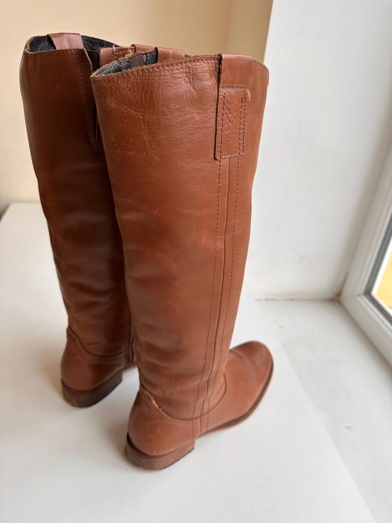 Pu&ograve; includere: Un paio di stivali alti in pelle marrone. Gli stivali hanno un design al ginocchio con punta arrotondata e tacco basso. La pelle ha una consistenza liscia e un colore marrone-rossastro caldo. Gli stivali sono in piedi.