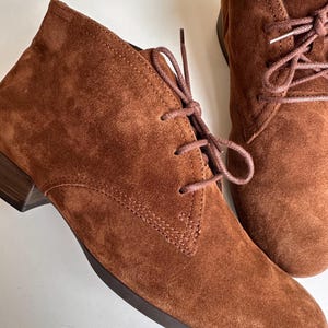 K&ouml;nnte beinhalten: Nahaufnahme von zwei braunen Wildleder-Ankle-Boots mit passenden Schn&uuml;rsenkeln. Die Stiefel haben einen kleinen Absatz und ein klassisches Chukka-Boot-Design. Das Wildleder hat ein weiches, strukturiertes Aussehen.
