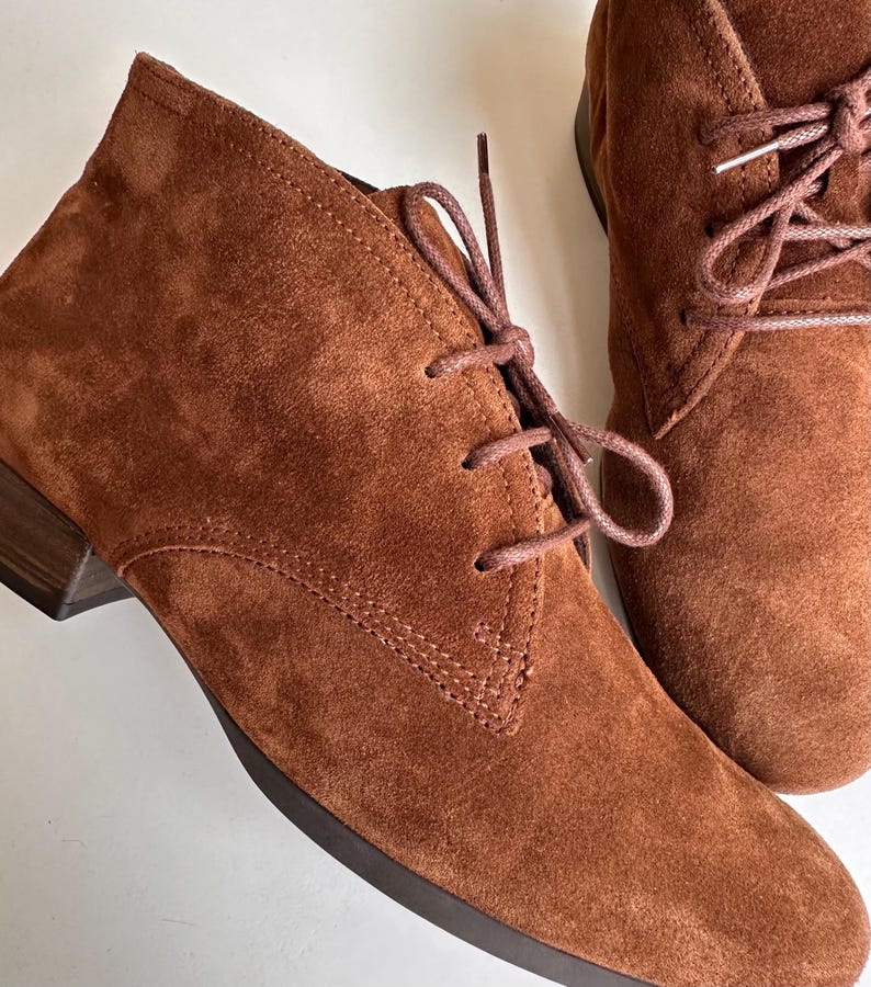 K&ouml;nnte beinhalten: Nahaufnahme von zwei braunen Wildleder-Ankle-Boots mit passenden Schn&uuml;rsenkeln. Die Stiefel haben einen kleinen Absatz und ein klassisches Chukka-Boot-Design. Das Wildleder hat ein weiches, strukturiertes Aussehen.