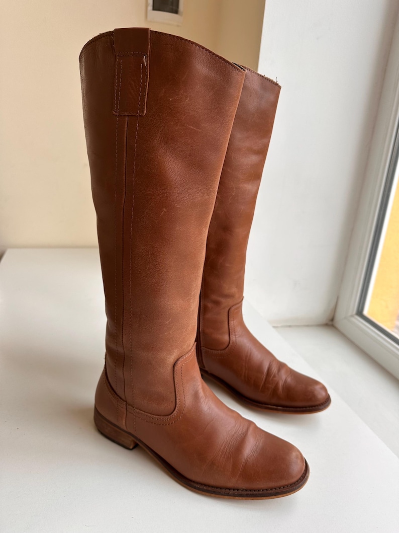 Pu&ograve; includere: Un paio di stivali alti in pelle marrone. Gli stivali hanno un design classico con punta arrotondata e tacco basso. La pelle ha un aspetto leggermente invecchiato, che ne aumenta il fascino vintage. Gli stivali sono adatti a varie occasioni.