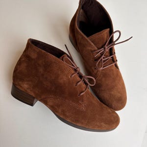 K&ouml;nnte beinhalten: Ein Paar braune Wildleder-Ankle Boots mit Schn&uuml;rung. Die Stiefel haben einen kleinen Absatz und eine runde Spitze. Die Schn&uuml;rsenkel und das Innenfutter sind in einem dunkleren Braunton gehalten. Die Stiefel stehen vor einem wei&szlig;en Hintergrund.