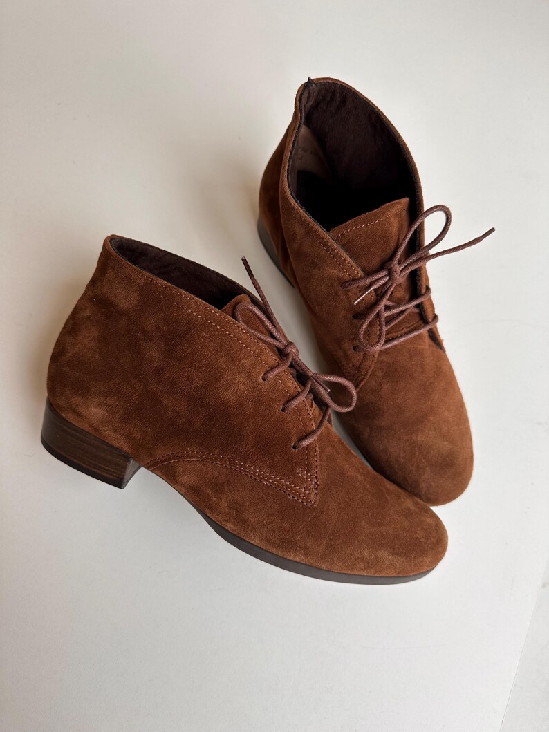 K&ouml;nnte beinhalten: Ein Paar braune Wildleder-Ankle Boots mit Schn&uuml;rung. Die Stiefel haben einen kleinen Absatz und eine runde Spitze. Die Schn&uuml;rsenkel und das Innenfutter sind in einem dunkleren Braunton gehalten. Die Stiefel stehen vor einem wei&szlig;en Hintergrund.