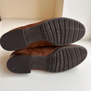 K&ouml;nnte beinhalten: Ein Paar braune Wildlederschuhe, Sohlen nach oben, mit dem Markennamen "Gabor". Die Sohlen sind dunkelbraun mit einem strukturierten Muster und den Worten "aircube". Die Schuhe haben eine warme braune Farbe.