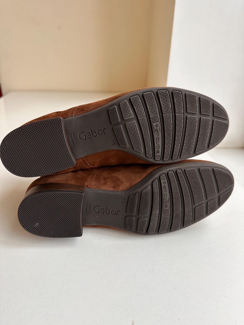 K&ouml;nnte beinhalten: Ein Paar braune Wildlederschuhe, Sohlen nach oben, mit dem Markennamen "Gabor". Die Sohlen sind dunkelbraun mit einem strukturierten Muster und den Worten "aircube". Die Schuhe haben eine warme braune Farbe.