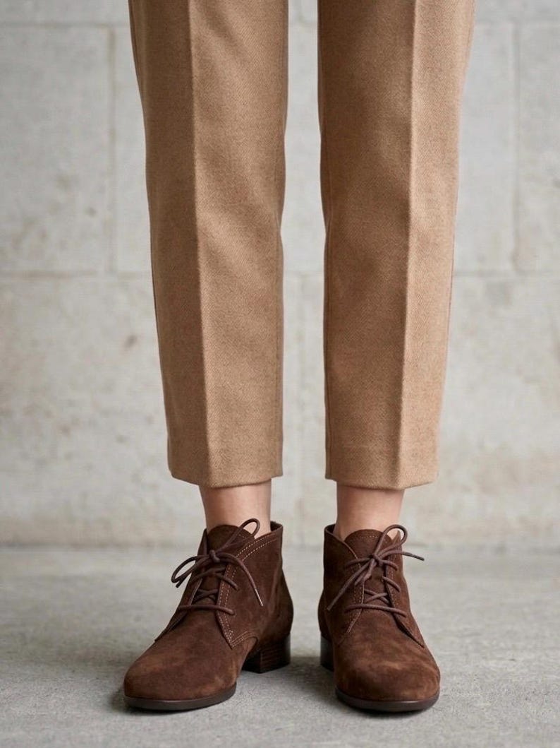 K&ouml;nnte beinhalten: Braune Wildleder-Ankle Boots mit Schn&uuml;rsenkeln, kombiniert mit beigefarbenen Hosen. Die Stiefel haben einen kleinen Absatz und ein klassisches Design, passend f&uuml;r den Alltag. Die Hose ist in einer neutralen Farbe gehalten.