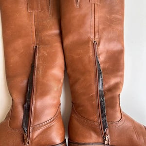Pu&ograve; includere: Un paio di stivali alti in pelle marrone con cerniere laterali. Gli stivali hanno un design classico con punta arrotondata e tacco basso. La pelle ha un aspetto leggermente invecchiato, che aggiunge al loro fascino vintage. Le cerniere sono marrone scuro.