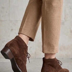 K&ouml;nnte beinhalten: Braune Wildleder-Ankle Boots mit niedrigem Absatz und Schn&uuml;rverschluss. Die Stiefel werden mit einer beigefarbenen Hose kombiniert. Das Schuhwerk hat ein klassisches Design, das f&uuml;r verschiedene Anl&auml;sse geeignet ist.