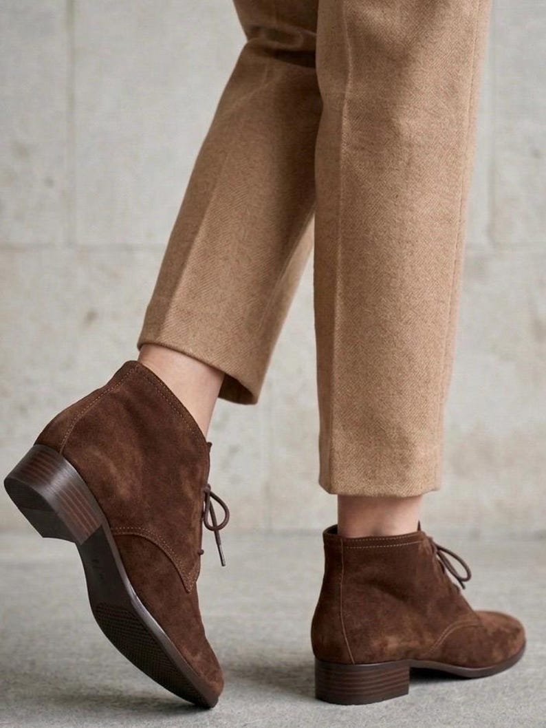K&ouml;nnte beinhalten: Braune Wildleder-Ankle Boots mit niedrigem Absatz und Schn&uuml;rverschluss. Die Stiefel werden mit einer beigefarbenen Hose kombiniert. Das Schuhwerk hat ein klassisches Design, das f&uuml;r verschiedene Anl&auml;sse geeignet ist.