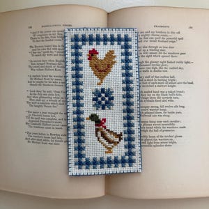 May include: A rectangular cross-stitch bookmark featuring a blue and white checkered border. The center design includes a brown chicken, a blue snowflake-like star, and a brown and white duck with a red neck detail, all stitched on white fabric.