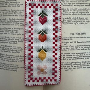 May include: A handmade cross-stitch bookmark with three embroidered fruits: a strawberry, an orange, and a lemon, plus a small flower. The bookmark has a red border and is set against an open book.