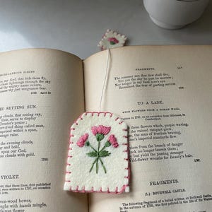 May include: A handmade, off-white felt tea bag with pink embroidered flowers and a pink stitched border. The tea bag rests on an open book with text and a teacup in the background. A small, matching felt tag is also visible.