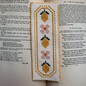 Puede incluir: Marcapáginas hecho a mano con punto de cruz con un patrón repetido de limones amarillos y flores rosas con hojas verdes, enmarcado por un borde dorado. El marcapáginas es rectangular y descansa sobre las páginas de un libro abierto.