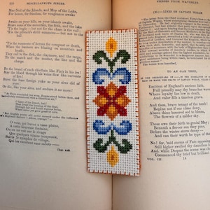 May include: A cross-stitch bookmark with a floral design in blue, red, green, and yellow. The bookmark is on a white background and is placed on an open book. The book is open to a page of text.