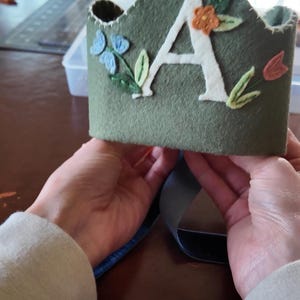 Op de afbeelding: Een groene viltkroon met een witte viltletter A en geborduurde bloemen in tinten blauw, oranje en roze.
