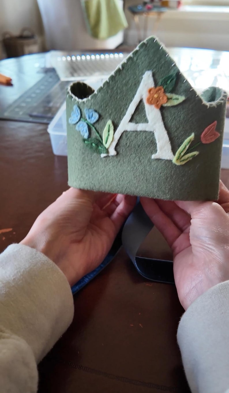 Op de afbeelding: Een groene viltkroon met een witte viltletter A en geborduurde bloemen in tinten blauw, oranje en roze.